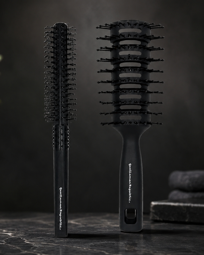 Two black hairbrushes displayed upright on a dark marble surface against a dark studio background, with a blurred plant and toiletry bottle in the background.