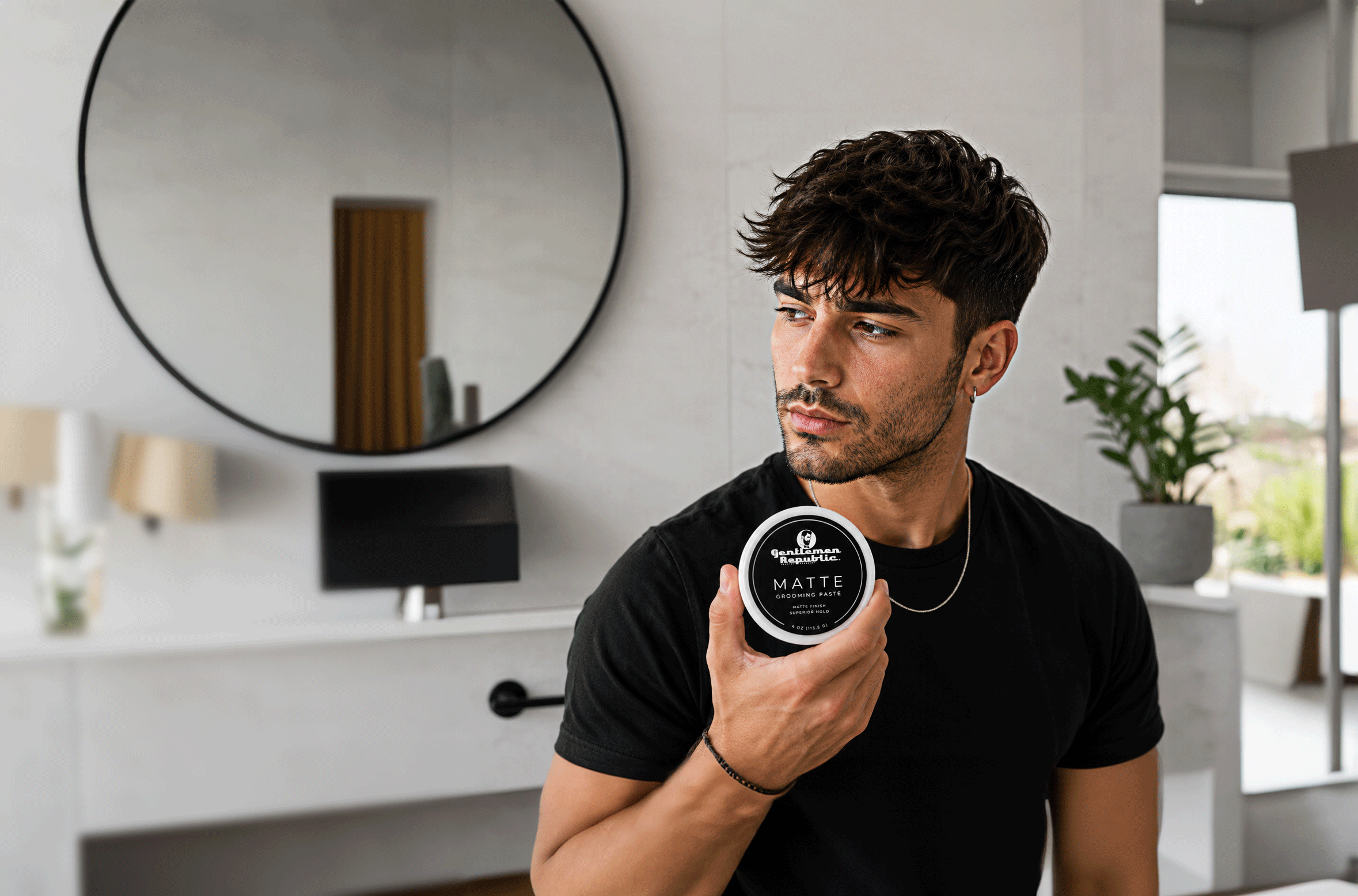 A man in a black t-shirt holds a round tin of 'Hair Artistry Matte' hair wax in a modern, bright bathroom setting.